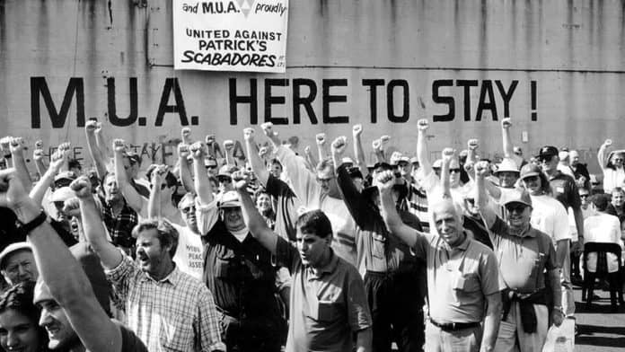 Black and white image of protest in Support of MUA. A Crowd of workers hold their fist in the air in front of a wall with the text "M.U.A. HERE TO STAY". A Poster hangs over the wall which says "United Against Patricks Scabadores"
