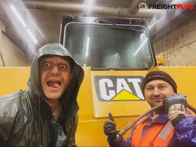 Two men covered in dirt, one wearing a khaki waterproof coat with a hood, the other wearing gloves, a beanie and an orange high-visibility vest pull faces in front of a CAT machine.