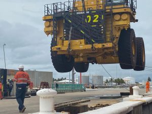 A man wearing an orange high-visibility long sleeved shirt with a black Freightplus logo stands in front of a large yellow dump truck hanging in the air being unloaded off a ship at a port on an overcast day. COVID-19 Updates for Australian Used Equipment Importers