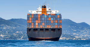 View of the back of a loaded container ship in blue ocean. Green hilly land is seen in the distant background. Canada joins investigation into Shipping Carriers