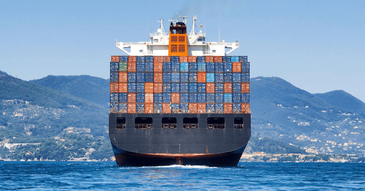 View of the back of a loaded container ship in blue ocean. Green hilly land is seen in the distant background