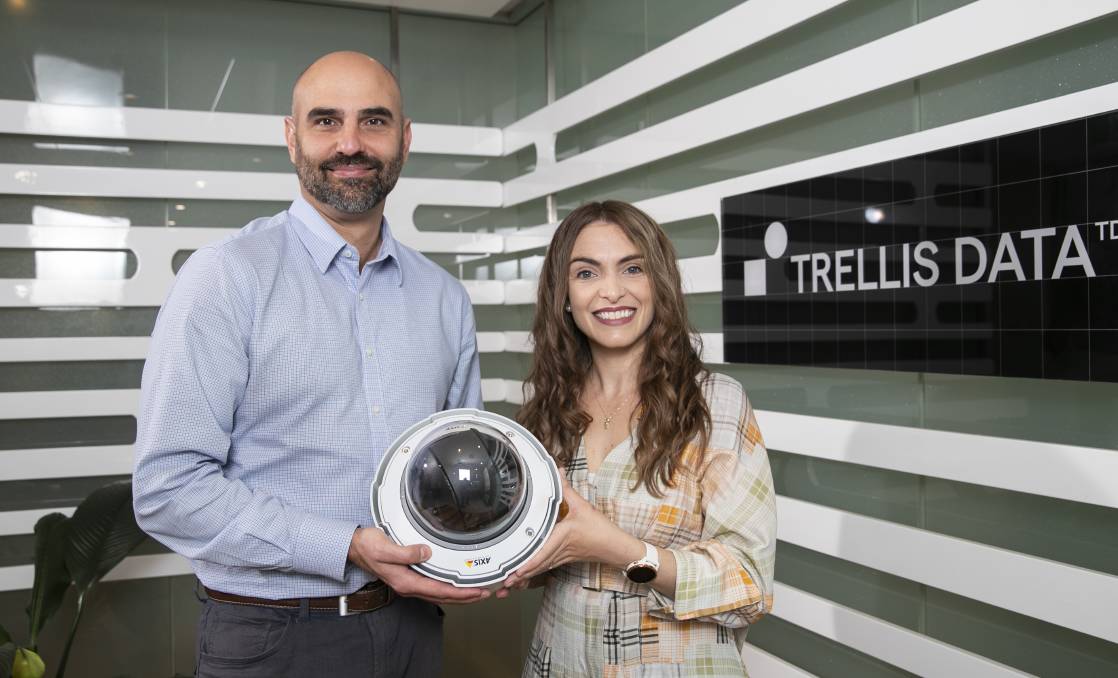 Man with bald head and facial hair in a blue shirt stands with woman with long brown hair and a floral dress hold a biosecurity camera in front of grey and white tripped wall with a black business sign with white text "Trellis Data"