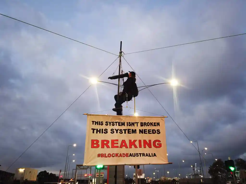 Person sits in a harness hanging from a large pole. Protesters disrupt port operations. From the top of the pole cables and ropes stretch out to secure it in place. The person is holding a large sign that reads "This system isn't broken, this system needs breaking #BlockadeAustralia". In the background two street lights are on against a dark cloudy sky.