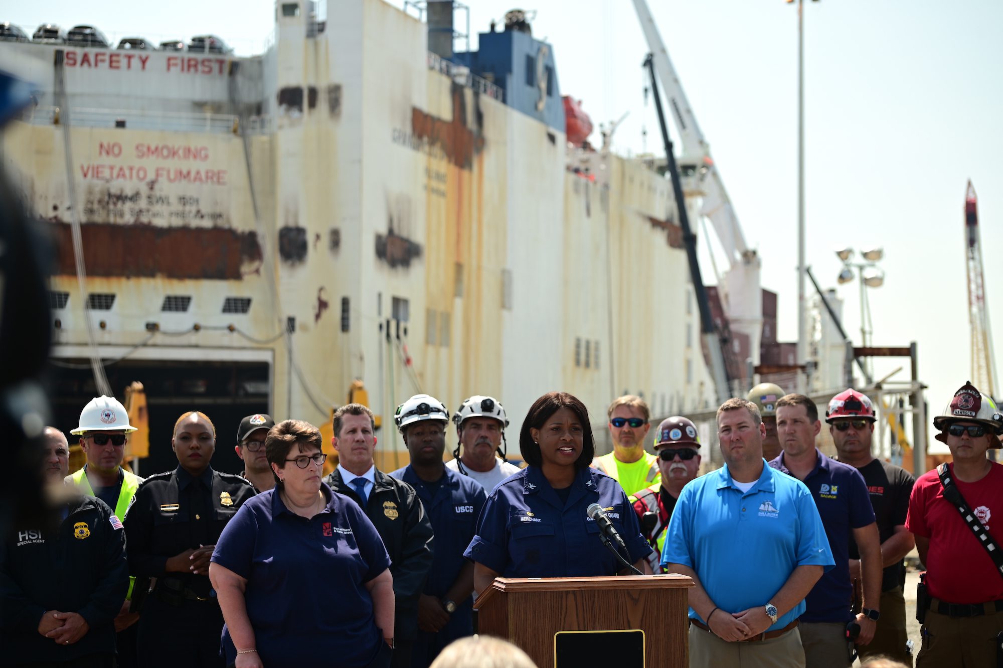 Emergency responders in a mix of coloured uniforms stand behind a podiem to gvive a press conferencee. In the backgrounds are the remains of the brurned RORO vessel