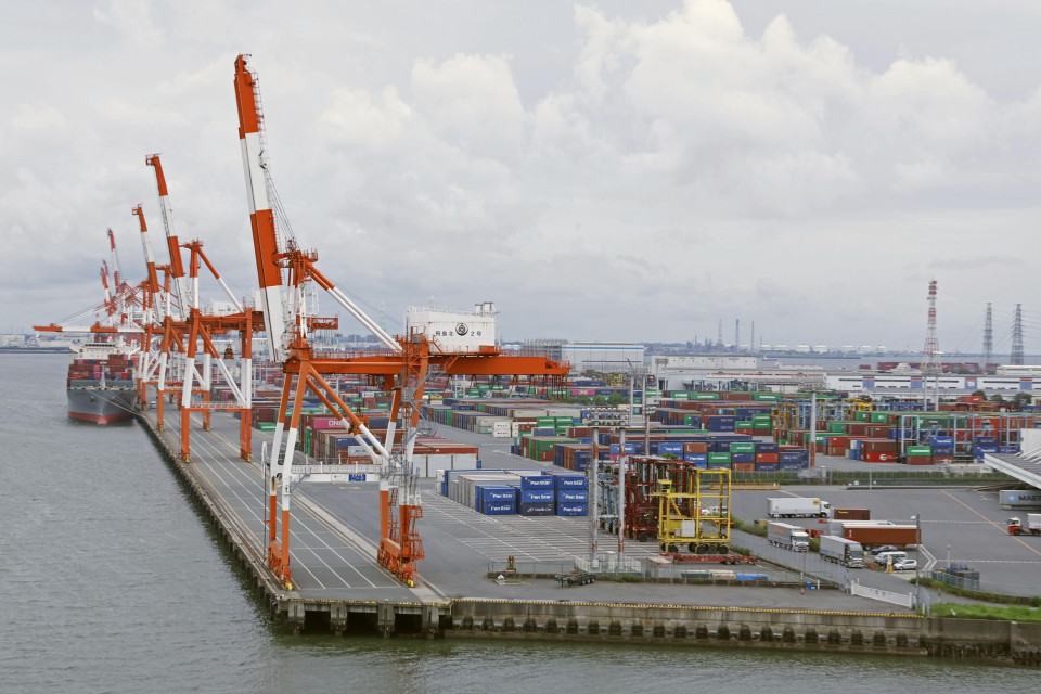 Orange and white cranes sit motionless on the waters edge at the port. In the background multicoloured containers sit. the sky is cloudy and water dark.