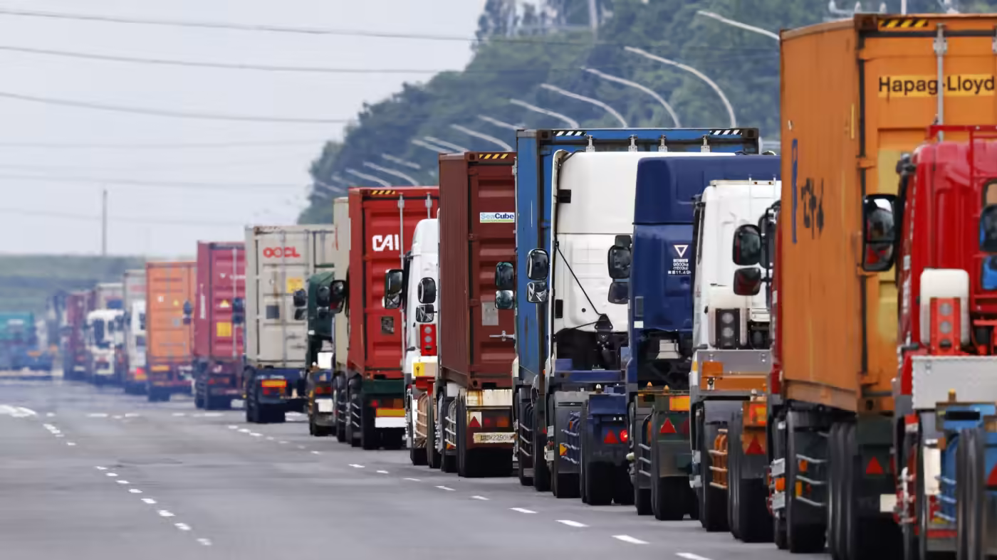 A large line of trucks, all with different coloured container on their trailers. The line of trucks goes as far as you can see. in the background there are lines of street lamps and trees