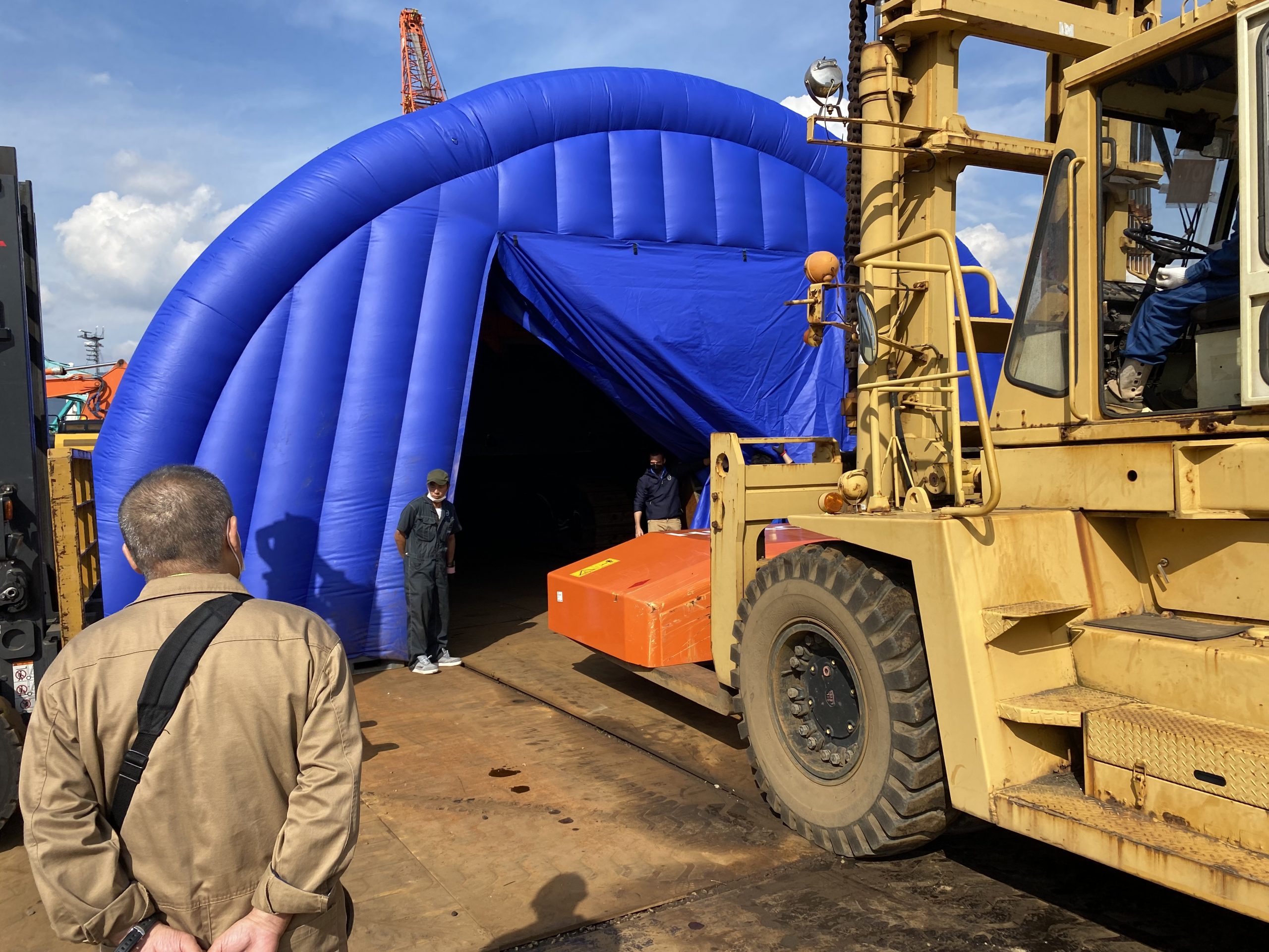 At hello forklift is carrying a bright orange piece of an Hitatchi ZX870 excavator into a bright blue fumigation tent. Two men dressed in boiler suits stand by the entrance to the tent. There is another man in the foreground in a tan long-sleeved shirt with black bag strap across his body. The sky is blue with a few white clouds. There are orange cranes in the background.