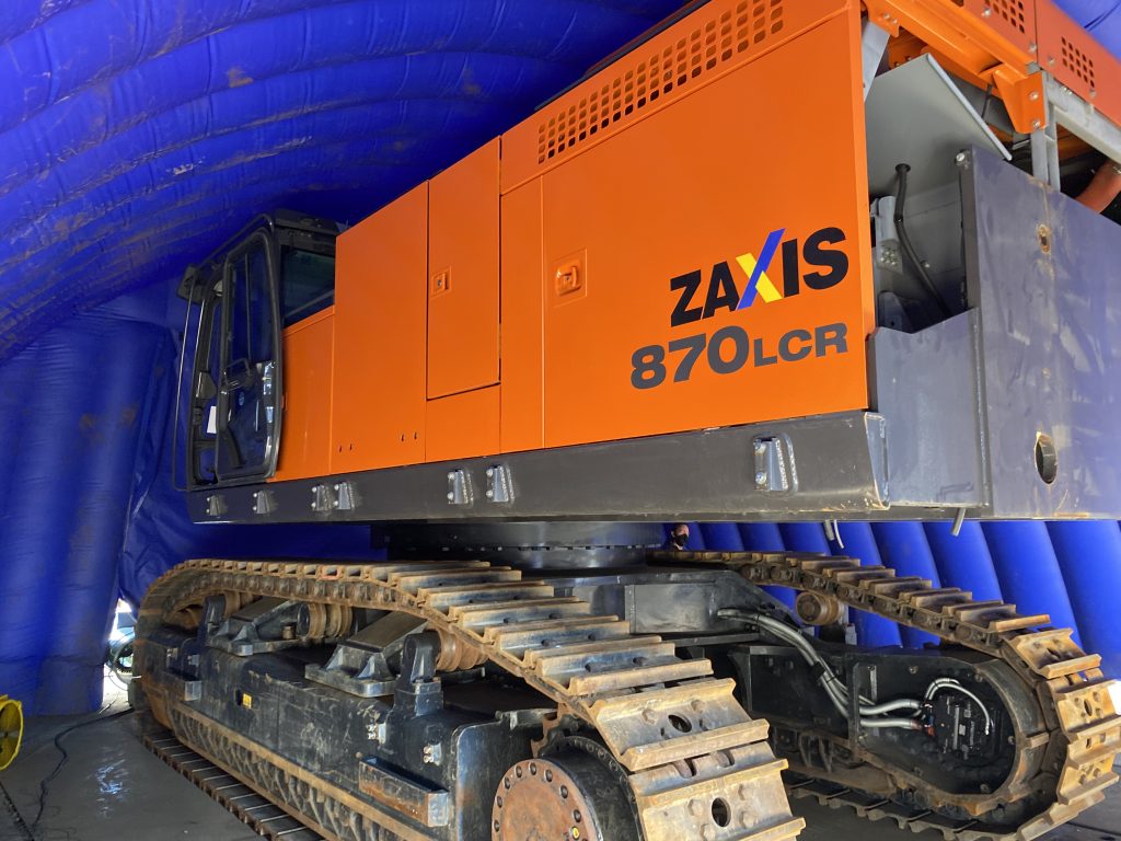 An orange Hitachi ZX870 excavator inside a blue fumigation tent receiving treatment for BMSB