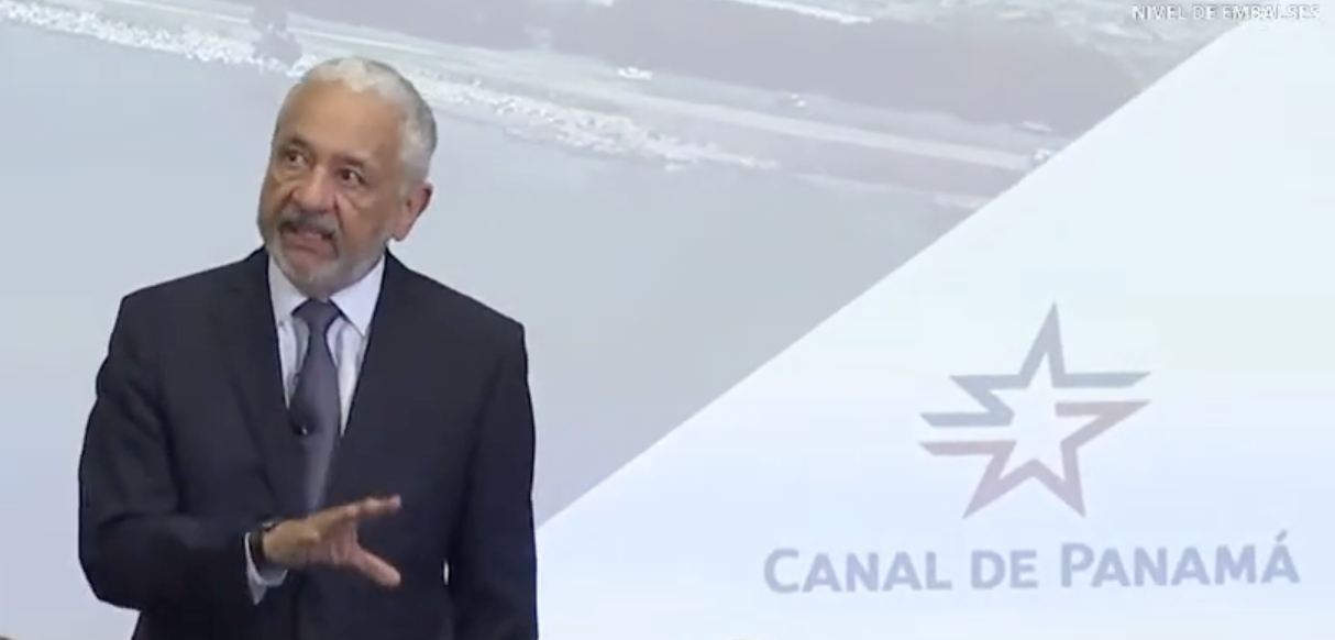 Middle ages business man with short grey hair in a dark suit stands in front of a projector screen which has a blue and red star logo with the text "Canal De Panama" under it.