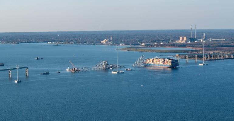 A wide shot showing the entire width of the river. What remains of the bridge jut out of the water. A loaded container ship rests at it's collision site