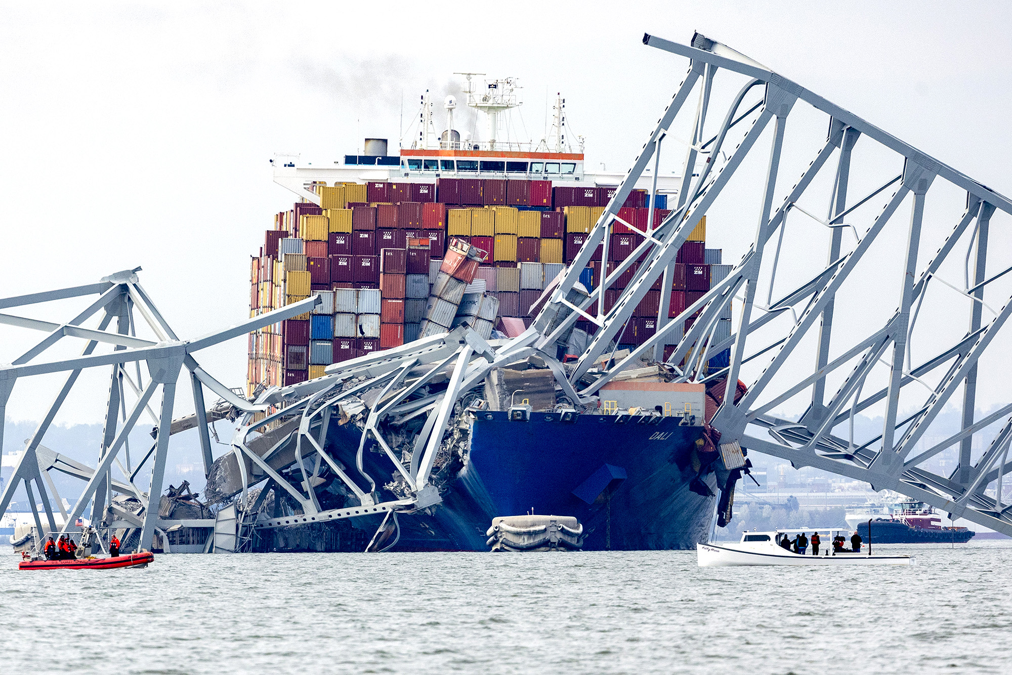 front on view of the MV Dali which the bridge collapsed on the bow of the ship. Multi-colour containers lean on an angle. smaller boats surround the ship.