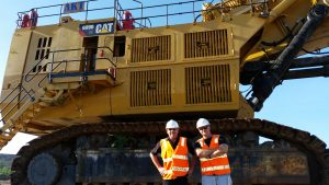 John Gleeson and Freightplus Japan country manager, Rod Mackay stand in orange hi-vis vests and white hard hats in from on a yellow CAT brand 6090 hydraulic mining shovel