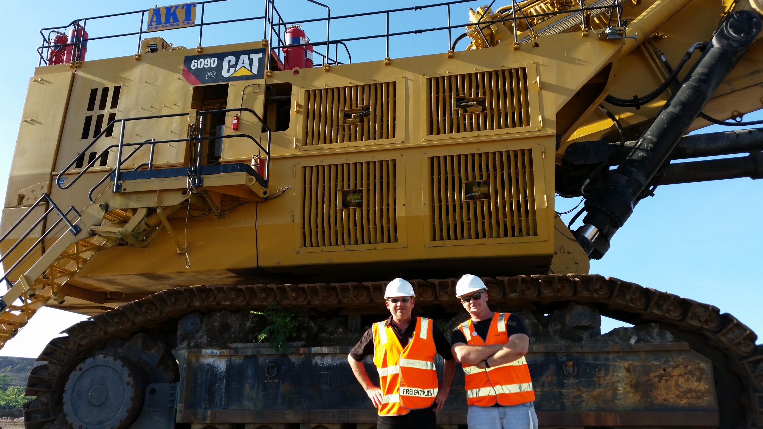 Rod and John stand in orange hi-vis vests and wait hard hats. Rod has his hands on his hips and John has his arms crossed. They stand in front of a large yellow CAT6090 excavator