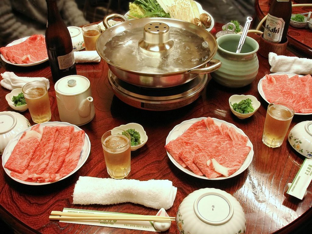 A japanese dinner table with a pot full o broth and vegetables in the centre. Plates of thinly sliced meat, napkins, chopstick, bowls, glasses of beer and Asahi bottles.