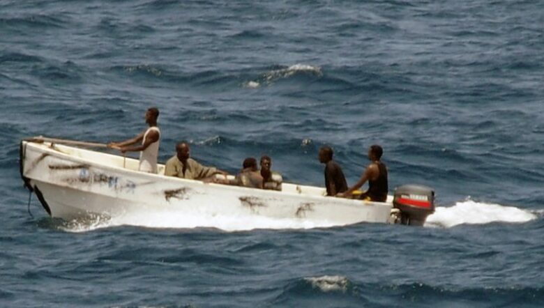 6 Somali pirates are in a white skiff in the oceaan. The skiff appears to have faded spay paint of faded black strips and a word written in blue paint. One man is standing at the bow holding onto a rope secured to the front go the boat. three men are sitting in the middle of the boat and two men sit up the back were one is driving the boat with a single Yamaha motor.