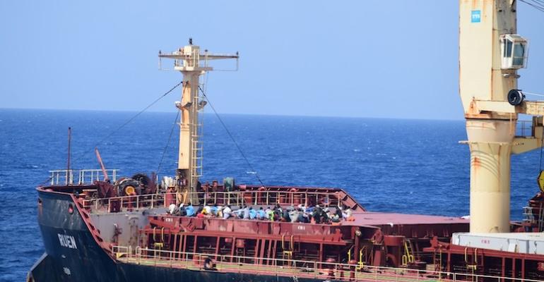 A group of Somali pirates sit atop the holds of a bulk carrier. The ship has yellow cranes, red interior and black exterior. The ship's name is visible on the side of the ship, written in white.