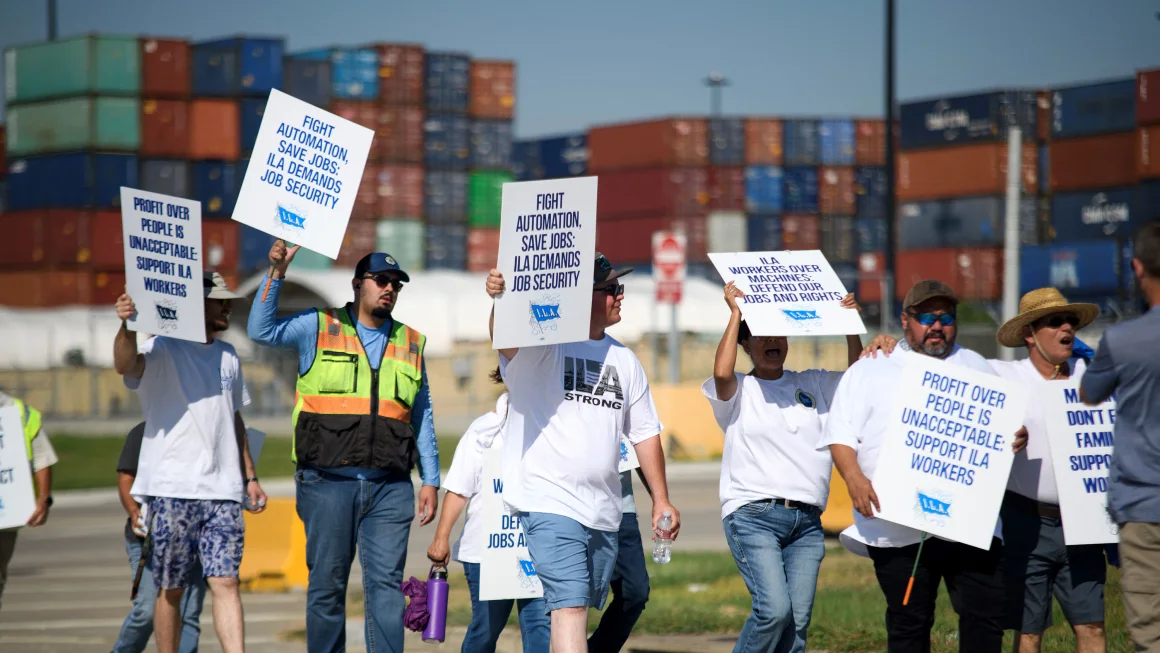 ILA Workers on strike at US port. Stacks of containers are in the background and the strikers hold signs.