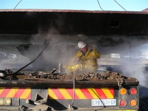 Steve Townsend wears a yellow worksuit and white hard hat, he's sitting on the back of a truck's flatbed trailer pressure washing the tray of a CAT brand 777 Dump truck that is lifted above him.