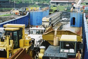 A blue landing craft, a barge-like vessel, docks on a muddy bank, it's been wet so you can see puddle in the mud. in the foregound are two CAT machines that have been loaded on first - one a dozer and they other a small articulated dump truck, behind them a road truck and at the back is a crane being loaded onto the LCT