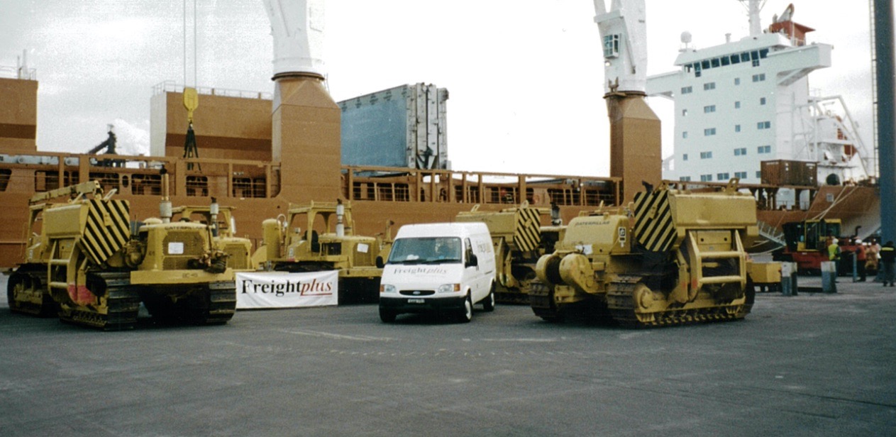 Yellow CAT brand Pipelayers sit at port waiting loading onto the breakbulk vessel docked in the background. Amongst the Pipelayers is a white Freightplus van and a banner with the 'Freightplus' original logo.