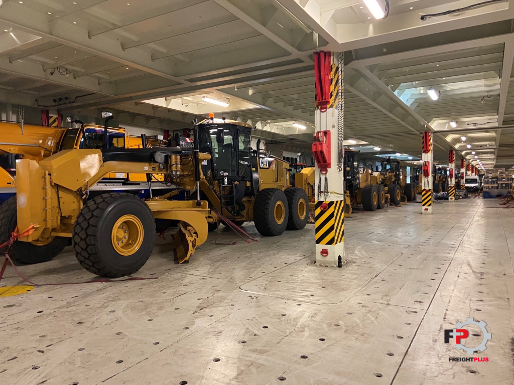 Inside the hull of a roll on roll off vessel, yellow caterpillar motor grader machines line up.