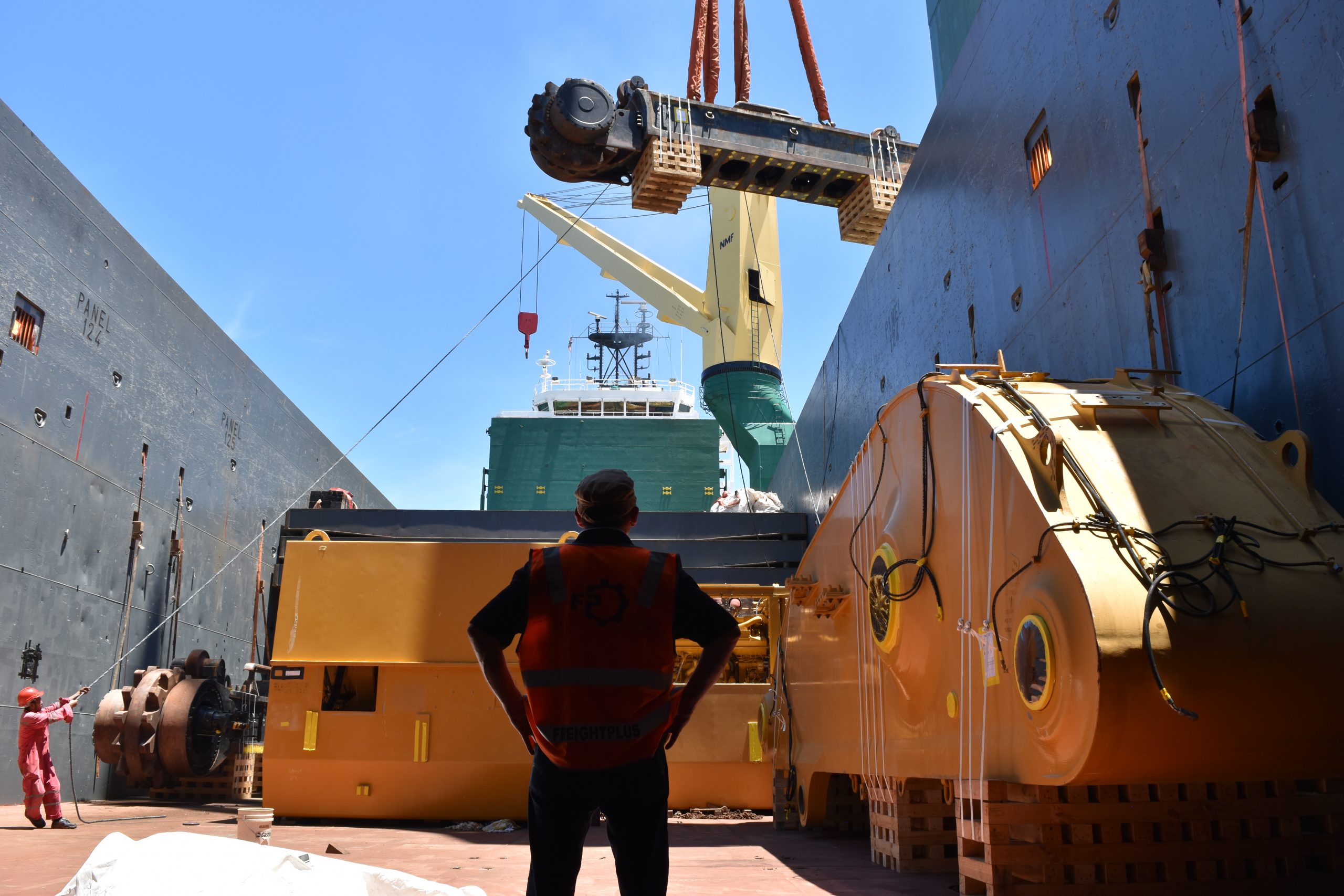 A silohetted man in a Freightplus branded orange hi-vis stands in the foreground of the image in the ship's hull. Large sections of a dismantled excavator are already loaded and another part is lifted in the air by the vessel's cranes to load in the hull